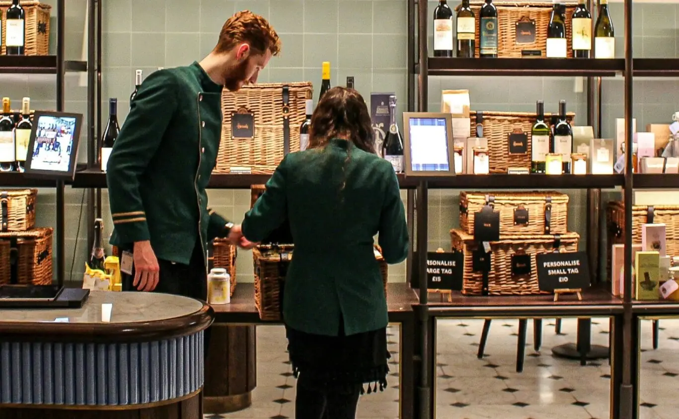 Two staff members inside Harrods with custom wine hampers and bottles of wine on the shelves behind them.